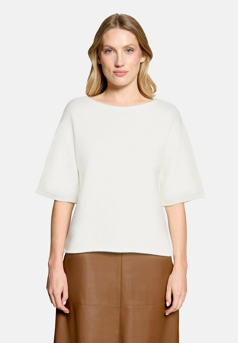 Femme aux cheveux blonds porte un haut blanc à manches courtes et une jupe en cuir marron, debout devant un fond clair uni.