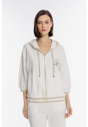 Kobieta w białej bluzie na suwak z złotą lamówką i detalami z kryształków, w połączeniu z dopasowanymi białymi spodniami, stojąca na jednolitym tle.