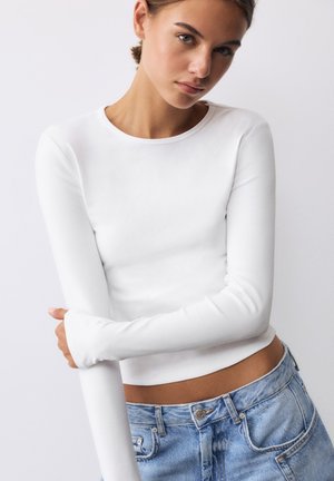 Jonge vrouw met een lichte huid die een aansluitend wit longsleeve topje en blauwe spijkerbroek draagt, poseert met gekruiste armen tegen een effen achtergrond.