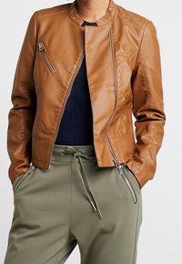 Hnědá kožená bunda s mírně přiléhavým střihem, mandarínským límcem a diagonálně umístěnými zipovými kapsami. Spárována se zelenými kalhotami.