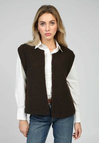 Gilet marron en tricot avec un motif texturé, devant ouvert et manches courtes, porté sur une chemise blanche à boutons et un jean bleu.