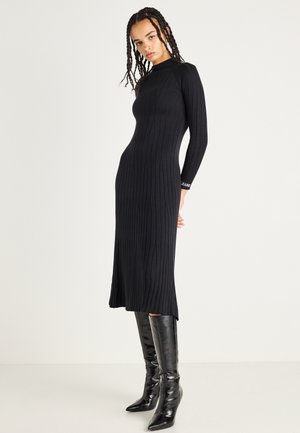 Rochie midi neagră cu volane, cu mâneci lungi, siluetă ajustată, guler rotund și slit pe lateral. Asortată cu cizme negre lucioase până la genunchi.