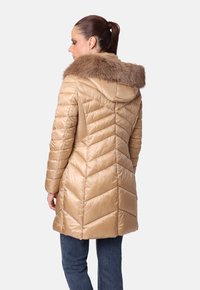 Mujer vistiendo un abrigo acolchado beige con capucha de piel, combinado con jeans azules, de espaldas contra un fondo liso.
