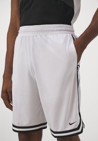 Shorts de sport blancs en tissu en maille, avec une ceinture noire et des rayures sur les côtés, portant un logo Nike sur l'ourlet inférieur.