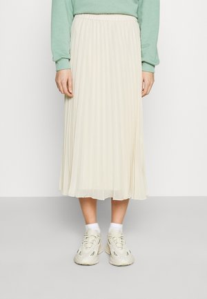 Jupe midi plissée en crème, en tissu léger ; dotée d'une taille élastique. Portée avec des baskets blanches et des chaussettes à la cheville.