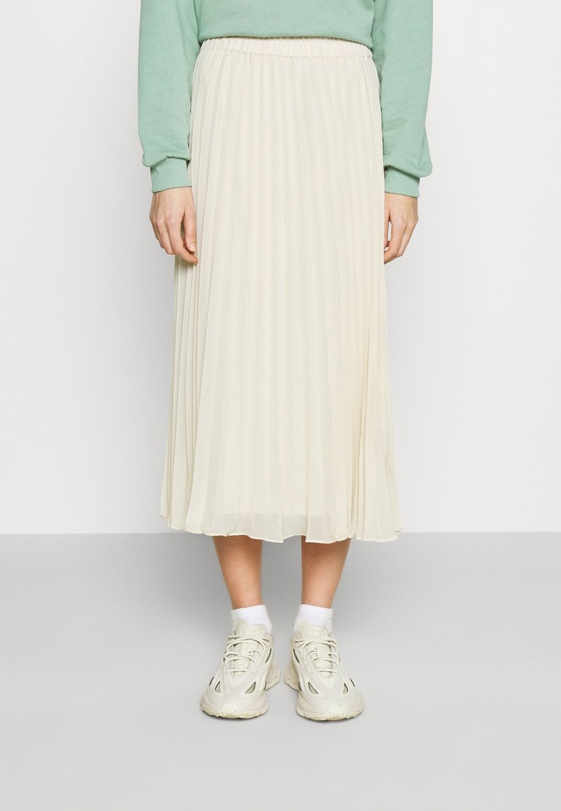 Gonna midi plissettata color crema, realizzata in tessuto leggero; presenta una vita elastica. Indossata con sneakers bianche e calzini alla caviglia.