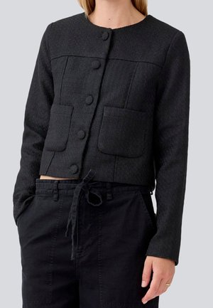 Vrouw draagt een zwarte getextureerde jas met knopen en twee zakken aan de voorkant, en zwarte broek met trekkoord, tegen een effen achtergrond.