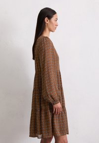 Braun gemustertes Kleid mit langen Ärmeln, einem sch tiered Rock und weißen Akzenten im Blätterdesign. Leichter Stoff mit fließender Silhouette.