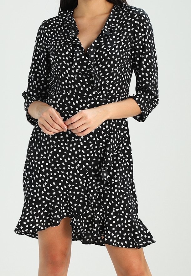 Femme portant une robe noire à pois blancs irréguliers, manches 3/4, encolure en V avec volants, et ourlet évasé, les mains jointes à la taille.