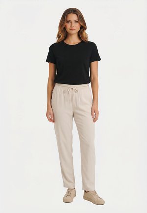 Donna in piedi che indossa una maglietta nera a maniche corte, pantaloni beige con coulisse e sneakers beige su uno sfondo bianco uniforme.