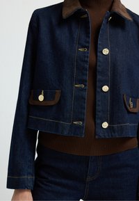 Tmavě modrá džínová bunda s hnědým semišovým límcem, zlatým prošíváním, dvěma předními kapsami a dekorativními knoflíky. Upevněný střih s dlouhými rukávy.