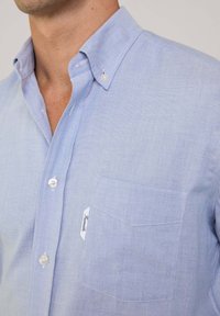 Camisa de botones de color azul claro hecha de tela texturizada, con un bolsillo en el pecho, detalles sutiles y botones blancos.