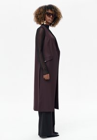 Długa bordowa kamizelka z bocznymi kieszeniami, noszona na czarnej bluzce z długim rękawem. Modelka ozdobiona dużymi okularami przeciwsłonecznymi i kwiatowymi kolczykami.