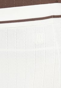 Λευκό πλεκτό πουλόβερ με σχέδιο καλωδίου, καφέ rib λαιμόκοψη και λευκή οριζόντια ρίγα. Περιλαμβάνει ανάγλυφο λογότυπο της μάρκας.