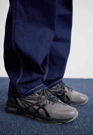 Osoba ubrana w ciemnoniebieskie szerokie dżinsy i szaro-czarne buty sportowe stojąca na niebieskim dywanie.