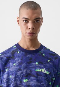 Blue short-sleeve shirt with a leaf pattern, featuring green accents and a small white logo. Crew neckline, textured fabric.
