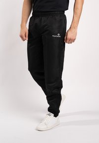Zwarte, lichte trainingsbroek met een elastische tailleband, voorzien van een klein wit logo op de dij, gedragen met witte sportschoenen.