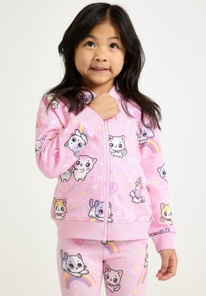 Mladá dívka ve svetru a kalhotách růžové barvy s potiskem kresleného kotěte a duhy, která mírně vzhlíží a usmívá se.