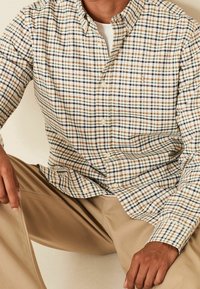 Licht crèmekleurig overhemd met een button-down kraag, voorzien van een multikleurig ruitpatroon in blauw, bruin en oranje. Kraag en logo op de borst zichtbaar.
