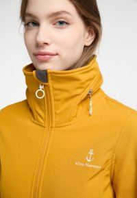 Giacca gialla impermeabile con alto colletto, dettaglio con zip e logo. Texture liscia con fodera grigia a contrasto e accessori decorativi.