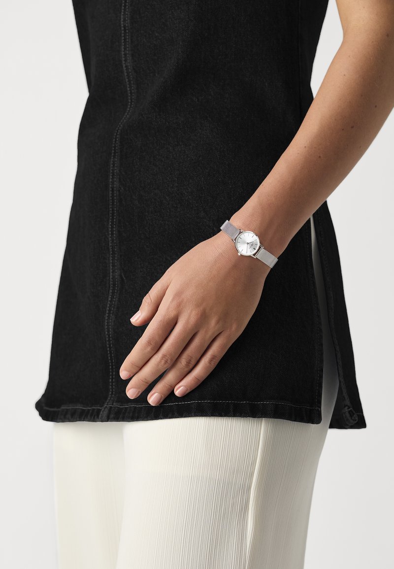 Orologio con cinturino in rete argentata, quadrante bianco, indicatori delle ore argentati e una cassa rotonda elegante, indossato su una mano contro un outfit in bianco e nero.