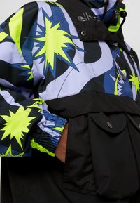 Veste avec motifs en étoile en néon jaune sur tissu bleu, noir et lavande, dotée de poignets élastiques et d'une poche avant boutonnée.