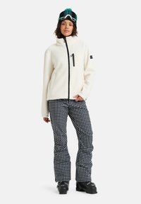 Fleece bunda v krémové barvě s černým zipem vpředu a kapsou. Pružné kalhoty v černobílé s kruhovými vzory. Černé boty a brýle.