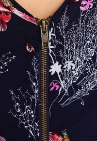 Navy fabric with floral print in white and pink; features a prominent gold zipper at the center, adding a textured accent.