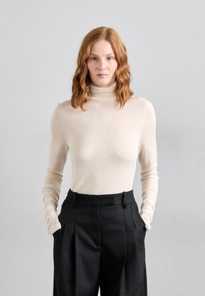 Suéter de cuello alto beige en una tela suave y ligera con mangas largas, combinado con pantalones negros de cintura alta con pliegues.