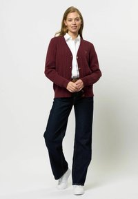 Burgundska pletena cardigan s rebrastimi manšeti in obodom, opremljen z mesingastimi gumbi. V kombinaciji s temnimi jeans hlačami in belimi športnimi copati.