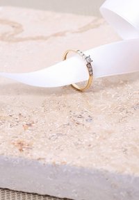 Gold ring featuring a central stone and three smaller stones on one side, resting on a textured stone surface with white satin ribbon.