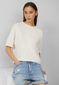 Krótki rękaw biały T-shirt wykonany z miękkiego materiału, cechujący się luźnym krojem, okrągłym dekoltem i subtelnymi podwiniętymi rękawami, noszony z postrzępionymi niebieskimi szortami.