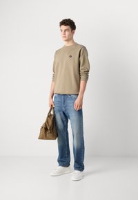 Khaki sweatshirt with a round neckline and small logo, paired with light blue straight-leg jeans and white sneakers, holding a brown bag.