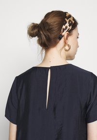 Frau mit braunen Haaren zu einem Dutt gebunden, die ein Stirnband mit Leopardenmuster, goldene Creolen und ein navyblaues Oberteil mit einem Rückenschlitz trägt.