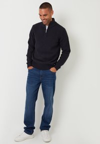 Marineblauer strukturierter Pullover mit halbem Reißverschluss, kombiniert mit blauen Jeans und weißen Sneakers, getragen von einem Model, das mit den Händen in den Taschen steht.