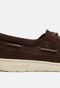 Bruine suède bootschoen met een witte rubberen zool, voorzien van subtiele logodetails en metalen veterogen. Klassiek ontwerp.