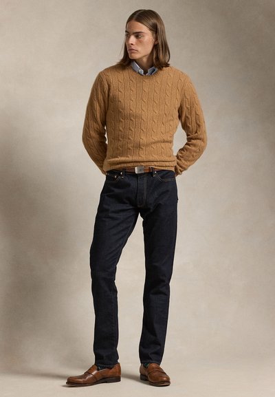 Muž nosící kamelí svetr s copánkovým vzorem, světle modrou košili s límečkem, tmavé džíny, hnědý pásek a hnědé kožené mokasíny, stojící se založenýma rukama za zády.