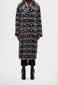 Cappotto lungo con motivo floreale nero, bianco e rosa. Presenta bottoni doppiopetto e una trama morbida. Indossato sopra pantaloni neri e stivali.