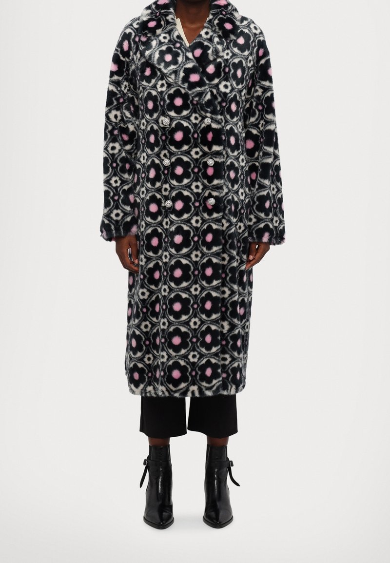 Cappotto lungo con motivo floreale nero, bianco e rosa. Presenta bottoni doppiopetto e una trama morbida. Indossato sopra pantaloni neri e stivali.