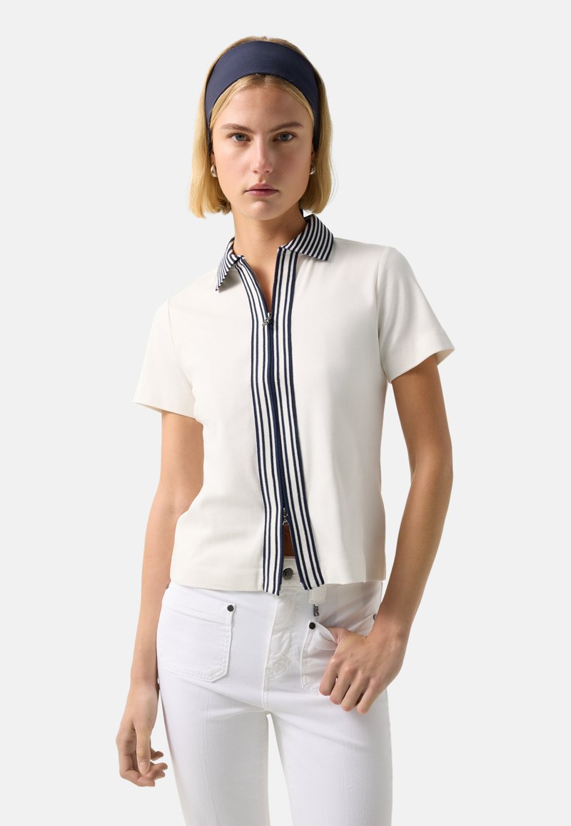 Woman wearing a white short-sleeve top with navy striped collar and zipper front, white pants, and a navy headband, standing against a plain background.