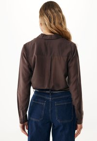 Donna con lunghi capelli mossi che indossa un blusa marrone a maniche lunghe infilata in jeans blu a vita alta con tasche posteriori visibili e una cintura a catena.