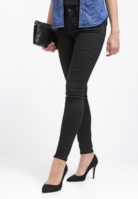 Jeans de mezclilla negros ajustados con una textura suave, con una cintura alta y diseño de cinco bolsillos. Combinados con tacones negros de punta y un bolso de mano negro.