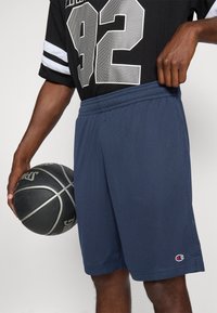 Dunkelblaue Mesh-Basketballshorts mit elastischem Bund, featuring ein Champion-Logo und einer lockeren Passform. Ein Basketball wird in einer Hand gehalten.