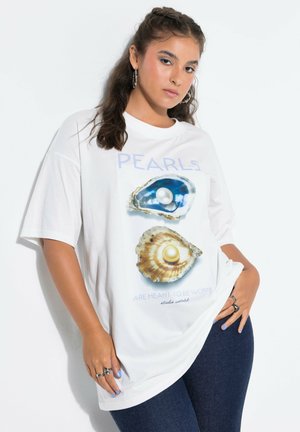 Young woman with braided hair wearing white oversized T-shirt with pearl shell prints and dark blue jeans, posing against white background.