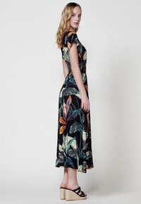 Maxi vestido negro con un patrón de hojas colorido, mangas cortas y una cintura ajustada. Lleva sandalias de cuña negras con tiras.