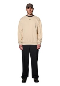 Beżowy sweter z prążkowaną strukturą i okrągłym dekoltem, z małym logo. W połączeniu z luźnymi czarnymi spodniami i teksturowanymi sneakersami, nosi czapkę.