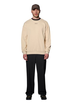 Beige sweatshirt met ribbeltextuur en ronde halslijn, voorzien van een klein logo. Gecombineerd met losse zwarte broek en textuursneakers, met een pet.
