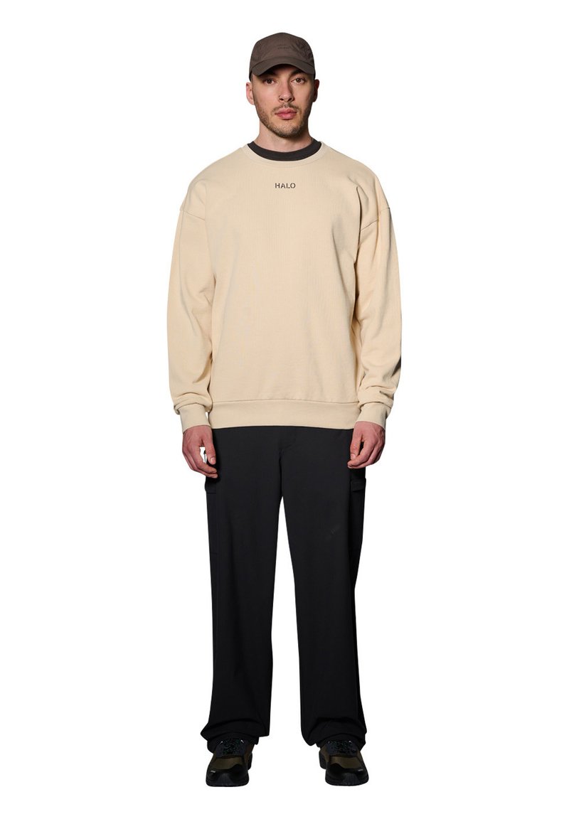 Beżowy sweter z prążkowaną strukturą i okrągłym dekoltem, z małym logo. W połączeniu z luźnymi czarnymi spodniami i teksturowanymi sneakersami, nosi czapkę.