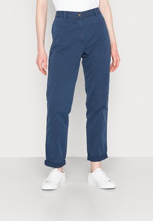 Osoba w granatowych spodniach podwiniętych do góry i białych trampkach, stojąca na jednolitym jasnym tle.