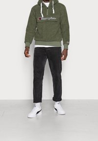 Maglione verde peloso con cappuccio, dotato di una tasca frontale e logo, abbinato a pantaloni neri a gamba affusolata e sneakers bianche con dettagli neri.